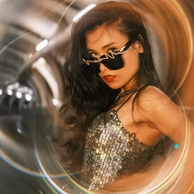 A stylish portrait of a woman in a sequined top and unique sunglasses, framed by swirling lights from a creative filter. The setting appears to be a modern, metallic tunnel, adding to the futuristic vibe.
