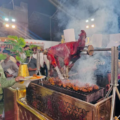 A whole tandoori goat roasting on a rotisserie, with smoke billowing from the grill. This is a spectacular centerpiece for any barbecue station.