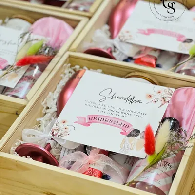 A personalized bridesmaid proposal hamper in a wooden crate. It's filled with thoughtful items like a custom wine glass, a bottle of sparkling rosé, a satin eye mask, and a card that perfectly pops the question.
