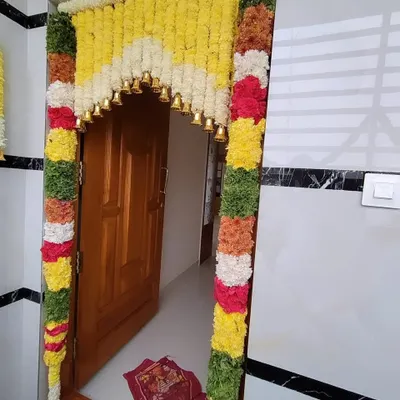 A vibrant and traditional entrance door decoration featuring a thick arch of yellow and white flowers, colorful side garlands, and auspicious brass bells.