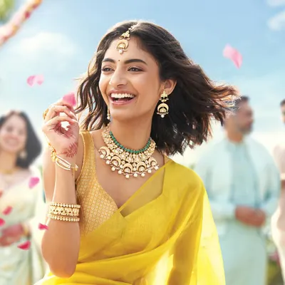 A candid, joyful moment from a Tanishq campaign, styling a modern Polki and emerald necklace set. The look is fresh and contemporary, pairing the jewellery with a simple yellow saree to appeal to a younger audience celebrating traditional events.