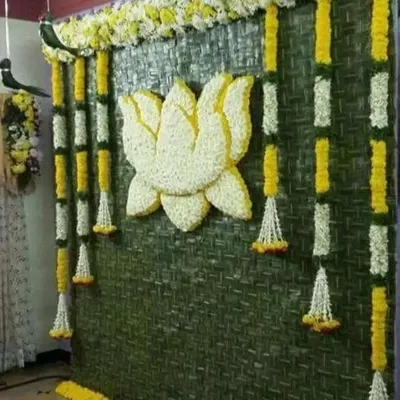 A beautiful Haldi backdrop made from a woven green mat, adorned with a lotus motif made of white petals and hanging garlands of yellow and white flowers.