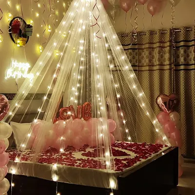 A dreamy birthday bedroom surprise featuring a sheer canopy illuminated with warm fairy lights. The decor includes soft pink and white balloons, rose petals arranged in a heart on the bed, and a "love" foil balloon to complete the romantic atmosphere.