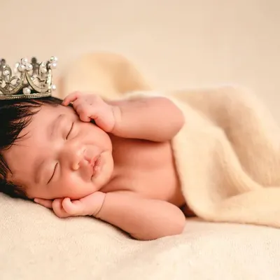 A classic "head on hands" pose on a soft, neutral blanket. This angle beautifully highlights the baby's delicate features and full head of hair, topped with a tiny crown.