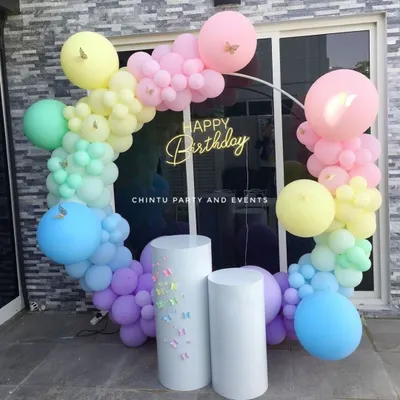 A pastel rainbow balloon ring decoration for a birthday. This outdoor setup includes a 'Happy Birthday' neon sign and white pedestals for the cake, creating a soft and dreamy photo backdrop.