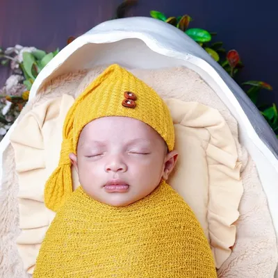 A close-up of a sleeping newborn in a yellow wrap and hat, nestled in a cozy prop. The focus is on the baby's peaceful face and tiny details.