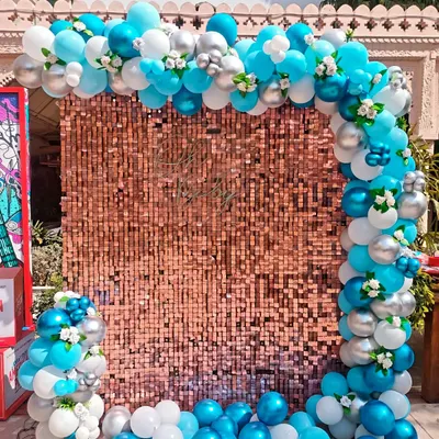 A breathtaking photo backdrop featuring a rose gold sequin wall. The wall is framed by a beautiful balloon arch in shades of turquoise, white, and silver, with delicate flower accents.
