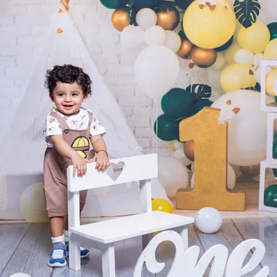 A happy toddler celebrating his first birthday. He stands proudly next to a "One" sign in a festive setup with a teepee and a beautiful balloon arch.