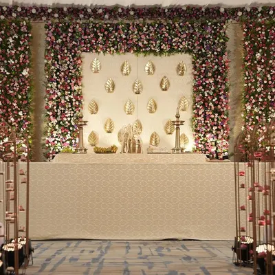 A graceful cream and gold stage for a South Indian indoor wedding. The backdrop features gold leaf motifs, framed by a dense wall of pink and purple flowers, with an aisle lit by candles and hanging lamps.
