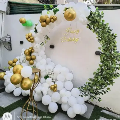 A beautiful and organic-style ring backdrop for an outdoor party. This design features a partial arch of white and gold balloons on one side and a cascade of artificial leaves on the other.