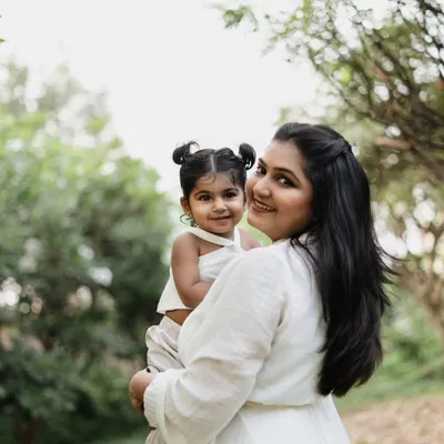 A mother holds her toddler daughter in a lush green garden. The natural light and beautiful surroundings create a soft and lovely portrait.