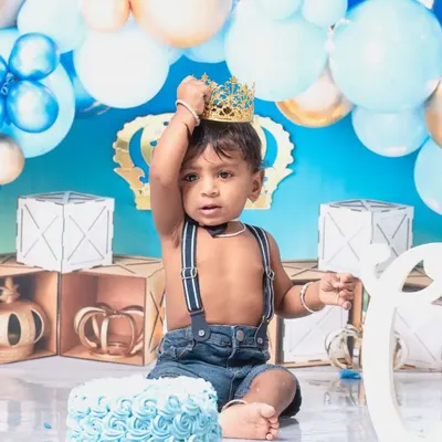 A little prince celebrating his first birthday with a royal blue cake smash. The setup includes a crown, balloons, and a delicious-looking cake ready to be smashed.
