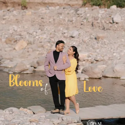 A sweet photo of a couple on a small wooden bridge over a stream, titled 'Blooms of Love', capturing a simple, happy moment.