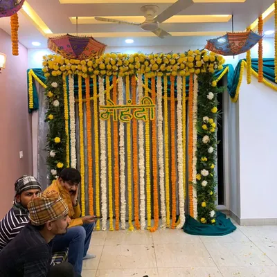 A classic Haldi and Mehndi backdrop featuring strings of fresh yellow and white flowers with a 'Mehndi' sign in Hindi. This setup is perfect for home functions and can be customized to fit your space.