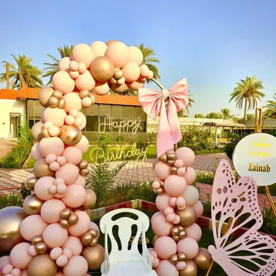 An outdoor butterfly-themed birthday setup with a beautiful pink and gold balloon arch. The decor includes a large pink butterfly prop, a 'Happy Birthday' neon sign, and a special chair for the birthday girl.