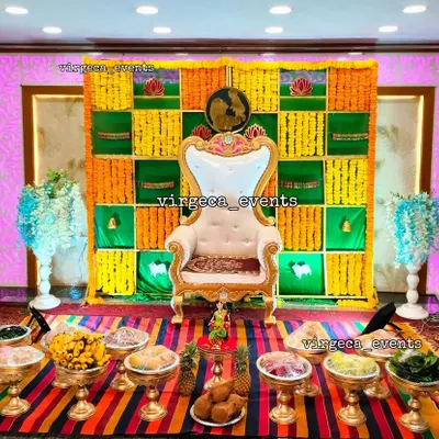 A vibrant Seemantha decoration featuring a traditional checkerboard backdrop of green, yellow, and orange marigolds. The setup is completed with an ornate chair and a full display of seer plates for the ceremony.