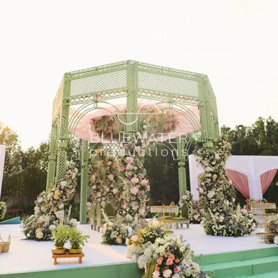 A full view of the French garden-inspired mandap, with its soft green lattice structure and abundant pastel florals creating a romantic focal point on the lush lawn.