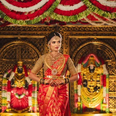 A bride standing before a magnificent temple-style mandap. The background features golden idols and is framed by rich red and white flower garlands, creating a divine atmosphere.