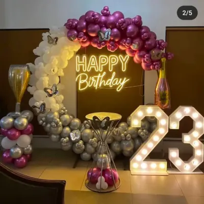A complete party setup for a 23rd birthday. It features a large circular balloon arch in purple, white, and silver, a custom "Happy Birthday" neon sign, and big light-up marquee numbers.