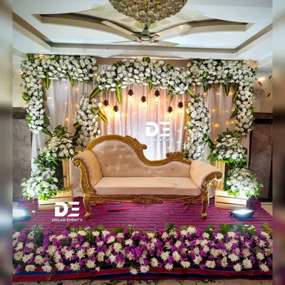 An elegant engagement or reception stage featuring a beautiful arch of white flowers, a classic chaise lounge, and warm accent lighting for a timeless look.