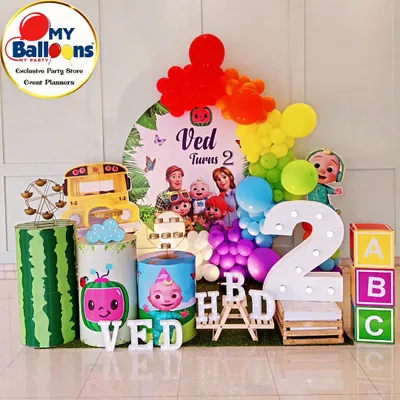 A complete Cocomelon-themed second birthday setup. This vibrant decor includes a rainbow balloon arch, the iconic yellow school bus, character-printed plinths, and a large light-up number '2'.