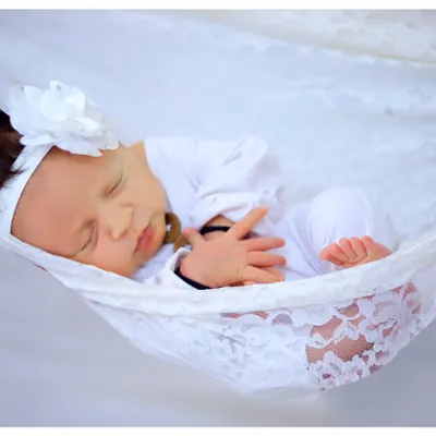 A peaceful newborn sleeps soundly in a white lace hammock against a clean white background. This style of portraiture emphasizes the baby's innocence and tranquility.