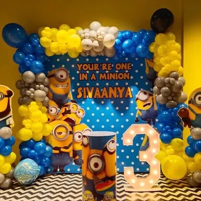 A complete Minions theme party setup for a 3rd birthday. This vibrant decoration features a "You're One in a Minion" backdrop, character cutouts, a light-up number '3', and balloon columns in the signature yellow and blue.