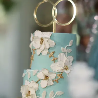 A close-up of this dreamy two-tiered engagement cake. The soft blue ganache is the perfect canvas for the handcrafted white and gold sugar flowers, topped with elegant interlocking rings.
