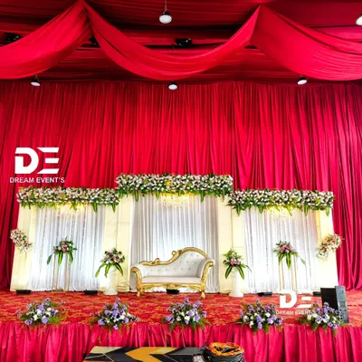 A classic reception stage featuring bold red ceiling drapery that creates a dramatic effect. The backdrop combines clean white panels with tiers of fresh flowers, perfectly framing the elegant couple's sofa.