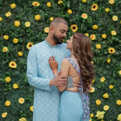 A sweet moment between a couple against a vibrant wall of yellow flowers during their Haldi ceremony. Their coordinated pastel blue outfits create a beautiful contrast, making for a fresh and modern portrait.