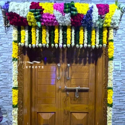 An elaborate main door flower decoration for a housewarming ceremony. This design features a dense frame of multi-colored flowers and jasmine buds, creating a grand and festive entrance.