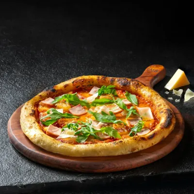 A classic ham and arugula pizza on a wooden board, shot with dramatic side lighting to highlight the crispy crust and fresh toppings.