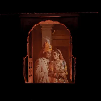 A cinematic, fairytale-like shot of the couple framed within a traditional Rajasthani jharokha window, creating a private and romantic moment.