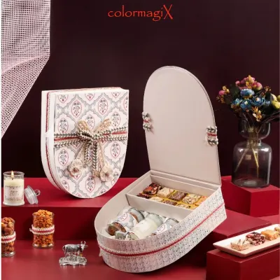 A uniquely shaped shield-style gift box with a traditional Indian motif and an elaborate woven ribbon. The box opens to reveal compartments filled with luxury sweets.