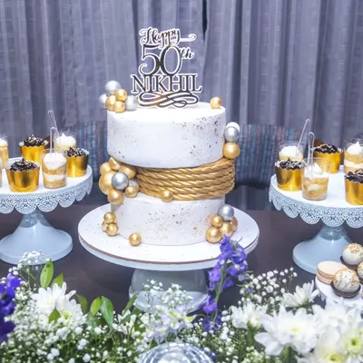A full view of the dessert table for a 50th birthday celebration. The elegant two-tier cake is flanked by dessert shots and macarons, all styled in a cohesive white, gold, and silver theme.