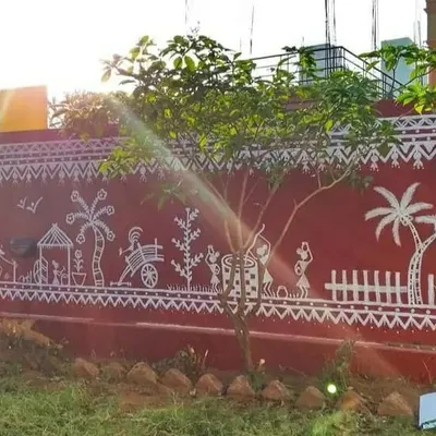 A wide view of the completed Warli mural, showcasing a detailed scene of rural life. This traditional art form, painted on a striking red exterior wall, tells a continuous story featuring people, animals, and nature.