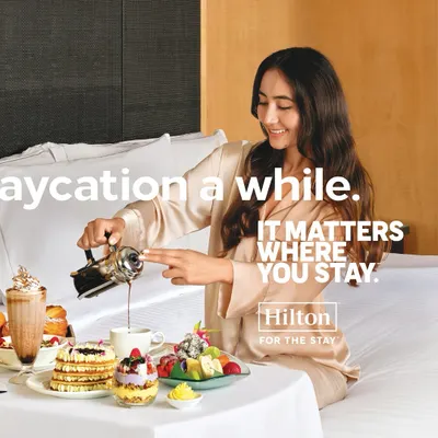 A serene moment styled for a Hilton Hotels campaign. The model's luxurious silk pajamas and the beautifully arranged breakfast tray create an aspirational 'staycation' scene, emphasizing comfort and indulgence.