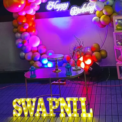 A surprise birthday party for a husband, featuring a modern ring circle balloon decoration. The setup includes a neon "Happy Birthday" sign, marquee letters, and a mix of pastel and chrome balloons for a stylish look.