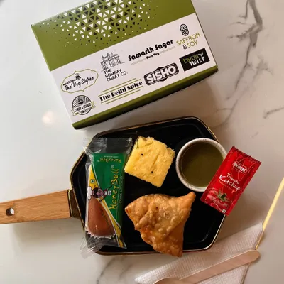 Our standard snack box is designed for convenience and taste. This one features a classic combination of a crispy samosa, soft dhokla, a sweet honey bell cake, and mint chutney, all packed in our branded, eco-friendly box.