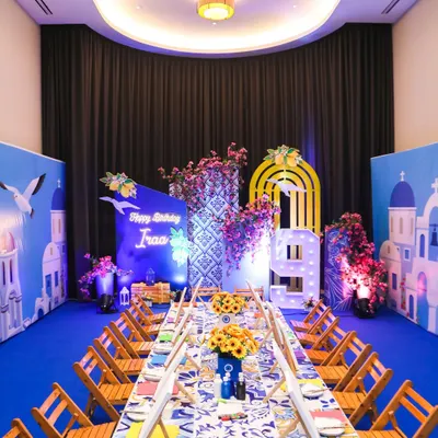 A stunning Greece-themed art party setup, with a long table ready for canvas painting, set against a backdrop of Santorini-style buildings and beautiful bougainvillea flowers.