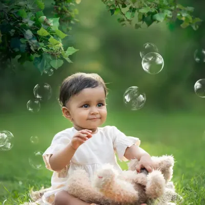 A baby sits on a blanket in a lush green park, mesmerized by floating bubbles. An outdoor sitter session like this is perfect for capturing natural wonder.