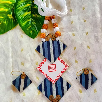 This geometric fabric jewelry set plays with shapes and patterns. The diamond-shaped pendant combines striped blue fabric with a hand-painted central element. The matching earrings and beaded cord complete this modern, lightweight accessory.