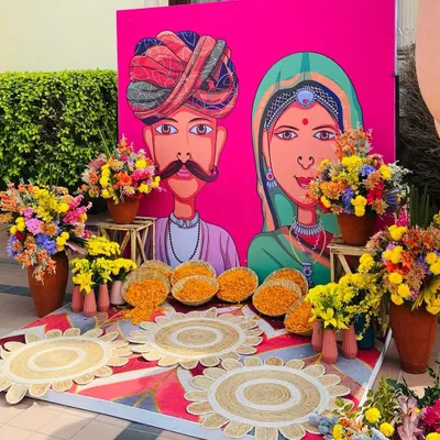 A quirky and colorful Mehendi photo booth with a custom Rajasthani-style caricature backdrop and vibrant floral arrangements.