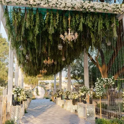 A truly grand and elegant wedding entrance. The walkway features a ceiling of hanging greenery and chandeliers, with the path lined by intricate white pedestals and floral arrangements.