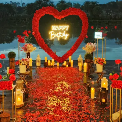 A stunning lakeside proposal setting at dusk. The scene is dominated by a large red floral heart arch and a thick carpet of red rose petals on a wooden deck, all surrounded by glowing lanterns and candles, reflecting beautifully on the water.