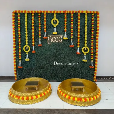 We designed this complete Haldi ceremony setup featuring a lush artificial grass wall backdrop, traditional marigold garlands, and a bright neon 'हळद' sign. The two large golden Urlis and matching patlas (stools) are also available from us on a rental basis.