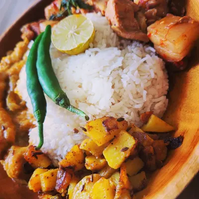 A hearty Naga meal plate centered around my signature Pork with Axone (fermented soybean). The plate also includes steamed rice, aloo fry, and a simple taro root curry, garnished with fresh green chili and lemon.