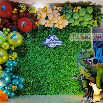 A close-up of a "Reyansh's World" Jurassic Park inspired theme. The setup includes a lush green wall and a dramatic balloon garland in red, yellow, and green.