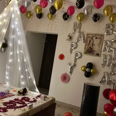 A complete romantic birthday surprise. This room decoration includes a bed canopy with fairy lights, rose petals on the bed, 'Happy Birthday' foil balloons on the wall, and balloons on the ceiling.