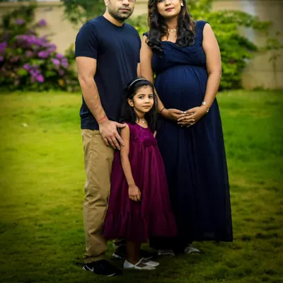 An expecting couple and their daughter pose for a beautiful outdoor maternity shoot in a lush green garden, capturing the family's anticipation in a natural setting.
